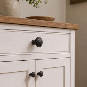 May include: A white wooden cabinet featuring a natural wood-toned top surface and three dark, matte-finished, circular knobs with a hammered texture. The cabinet includes one upper drawer and two lower doors, each accented with these round metal knobs.