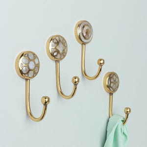 May include: Four brass wall hooks with mother-of-pearl inlay. Each hook features a different design, including a honeycomb, starburst, and concentric circle pattern. One hook holds a light green scarf. The hooks are mounted on a light blue wall.