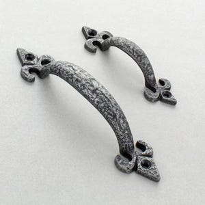 Op de afbeelding: Twee handgrepen voor kasten in antieke stijl met een getextureerde, gevlekte grijze afwerking. Elke handgreep heeft een gebogen stang en decoratieve fleur-de-lis uiteinden, die een vleugje vintage charme toevoegen.