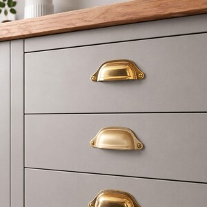 Peut inclure: Trois poignées de tiroir en forme de coquille en laiton poli fixées sur un meuble gris clair à trois tiroirs horizontaux. Chaque poignée présente un design semi-circulaire avec deux vis visibles sur les brides de fixation, offrant un contraste métallique sur la surface grise plane des façades de tiroirs.