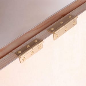 May include: Two rectangular brass hinges with a brushed finish, each featuring three circular screw holes with flathead screws installed. The hinges are mounted onto the edge of a light-colored wooden panel, showing a metallic gold tone.