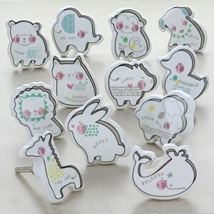 May include: A collection of white ceramic cabinet knobs in various animal shapes, including a bear, elephant, monkey, dog, cat, hippo, duck, lion, rabbit, sheep, giraffe, and whale. Each knob features a unique design with pastel colors and text.