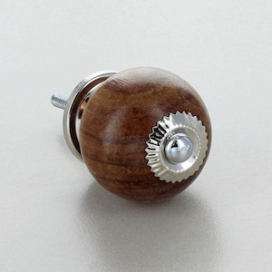 May include: A round wooden knob with a silver metal trim and a small silver metal button in the centre. The knob is brown with a wood grain pattern.
