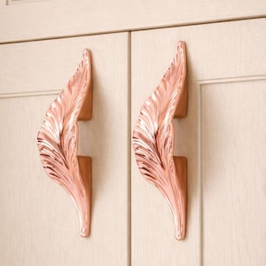 May include: Two rose gold cabinet pulls in a feather design. The pulls are mounted on a light-coloured cabinet door. The feather design has detailed veining and a curved shape, adding a decorative touch to the cabinetry.