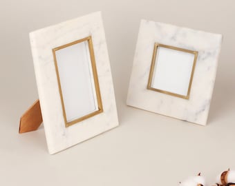 May include: Two white marble picture frames with gold-colored metal borders. One frame is rectangular and propped up with a wooden stand. The other frame is square. Both frames have a clean, minimalist design.