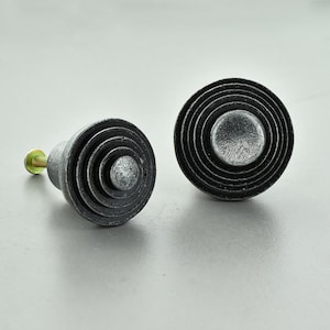 May include: Two cabinet knobs with a concentric circle design. The knobs are dark grey with a distressed finish, and each has a small, silver-coloured centre. One knob has a gold-coloured screw attached.