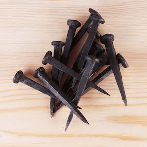 May include: A collection of dark gray, hand-forged iron nails with square shanks and flat, irregular heads. These metal fasteners feature a textured, matte finish and pointed tips, arranged in a scattered pile to show their individual shapes.