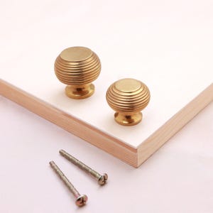 May include: Two round, gold-coloured metal cabinet knobs featuring a horizontal ribbed texture and a smooth, flat top surface. The knobs are accompanied by two matching metal mounting screws with threaded shafts, designed for furniture installation.
