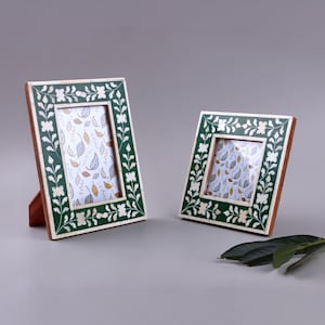 Op de afbeelding: Twee decoratieve fotolijsten. De lijsten hebben een donkergroene rand met een bloemmotief en een lichtgekleurde inleg. De lijsten zijn rechthoekig en hebben een houten achterkant. De foto's in de lijsten hebben een bladerenpatroon.