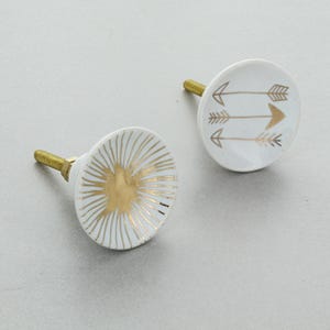May include: Two white ceramic cabinet knobs with gold detailing. One knob has a sunburst pattern, the other features three gold arrows. Each knob has a gold-coloured screw. These decorative knobs are suitable for adding a stylish touch to furniture.