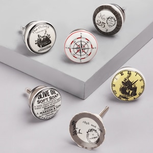 May include: Five ceramic drawer knobs with different designs. The knobs are white with black and red accents. The designs include a compass, a clock, a typewriter, a bottle of soap, and a stamp.