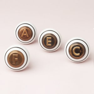 Peut inclure: Quatre boutons en céramique blanche avec une bordure noire et des accents de lettres dorées. Les lettres sont A, E, C et F.
