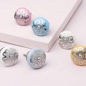 Pode incluir: Seis puxadores de armário de cerâmica coloridos. Os puxadores são em tons de rosa, azul, branco, amarelo e azul claro. Cada puxador tem um design floral prateado. Os puxadores estão sobre uma superfície rosa claro.