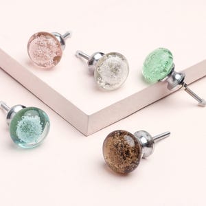 May include: Five decorative glass cabinet knobs in various colors: pink, white, green, teal, and brown. Each knob features a bubble-like design within the glass and a silver metal base and screw.