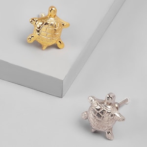 Könnte beinhalten: Zwei dekorative schildkrötenförmige Schrankknöpfe. Einer ist goldfarben, der andere silberfarben. Die Knöpfe haben ein strukturiertes Schalendesign und sind auf weißem Hintergrund montiert.