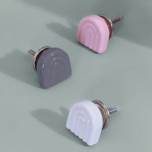 May include: Three ceramic cabinet knobs in a rainbow design. One is grey, one is pink, and one is white. Each knob has a silver metal base and screw. The knobs are arranged on a light green surface.