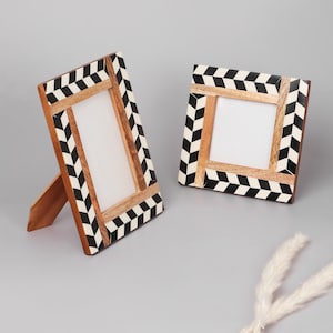 May include: Two decorative wooden picture frames. The frames have a natural wood interior and a black and white geometric pattern on the border. One frame is propped up with a wooden stand, while the other lies flat. The frames are on a gray surface.