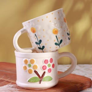 Peut inclure: Deux tasses en céramique blanches empilées. La tasse du dessus présente un motif floral avec des accents jaunes et verts. La tasse du dessous présente un motif floral similaire avec des détails jaunes, roses et verts. Les deux tasses ont des anses incurvées.