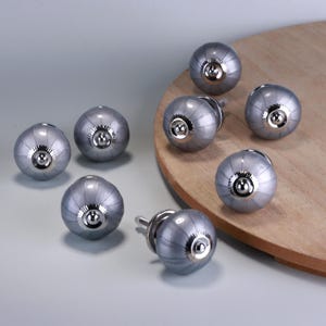 May include: Eight grey and silver cabinet knobs are displayed. Each knob features a rounded, ribbed design with a silver centre and a mounting screw. The knobs are arranged on a light grey surface and a wooden board.