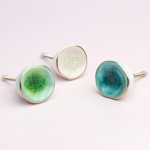 May include: Three decorative cabinet knobs with a unique, organic design. Each knob features a round, glazed ceramic face in shades of green, white, and teal, accented with gold trim. The knobs have a white base and a silver screw.