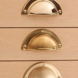 Op de afbeelding: Drie halfronde komgrepen voor lades gemonteerd op lichtgekleurd hout. De bovenste greep is van gepolijst messing, de middelste van verouderd messing met patina en de onderste heeft een matte satijnen messing afwerking. Elke greep is bevestigd met drie schroeven.