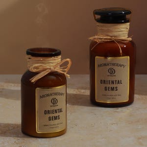 May include: Two brown glass aromatherapy candles with black lids and raffia accents. The labels read "Aromatherapy Oriental Gems" and "Hand Poured Soy Wax". One candle is lit, with smoke rising.
