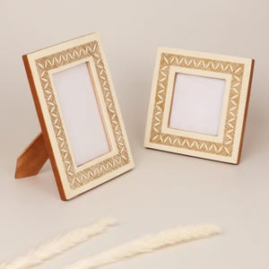 May include: Two decorative photo frames. One frame is rectangular and propped up with a wooden stand. The other frame is square. Both frames have a cream-colored border with a detailed, repeating pattern and a brown wooden frame.