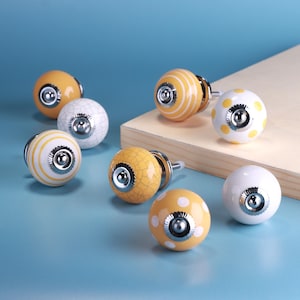May include: A selection of decorative ceramic cabinet knobs in various designs. The knobs feature patterns such as stripes, polka dots, and crackle textures in yellow, white, and orange hues. Each knob has a silver-toned metal base.