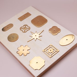 May include: A collection of eleven brass-colored metal plates in various geometric and organic shapes, including stars, circles, squares, and clover-like forms. Each piece features a single centered hole for mounting or attachment.