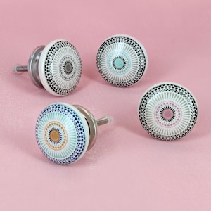 May include: Four decorative ceramic cabinet knobs with a white base and colorful geometric patterns. Each knob features a different color scheme, including blue, teal, pink, and gray, with a black dotted border. The knobs have silver metal hardware.