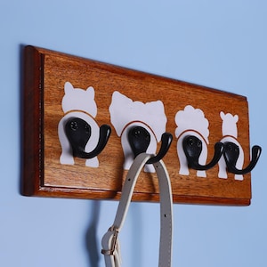 Può includere: Appendiabiti da parete in legno con quattro ganci in metallo nero. Il portabiti presenta ritagli bianchi a forma di animale. Una tracolla di borsa di colore chiaro è appesa a uno dei ganci. Il legno ha una calda finitura marrone rossiccio.