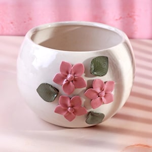 May include: A white ceramic plant pot with a rounded shape. The pot is decorated with three pink flowers and three green leaves. The pot is on a pink surface.