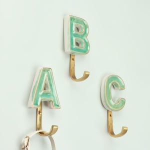 Peut inclure: Trois patères murales décoratives en forme de lettres A, B et C. Chaque lettre est blanche avec un glaçage turquoise et une bordure dorée, fixée à un crochet en laiton. Parfaites pour suspendre des objets.