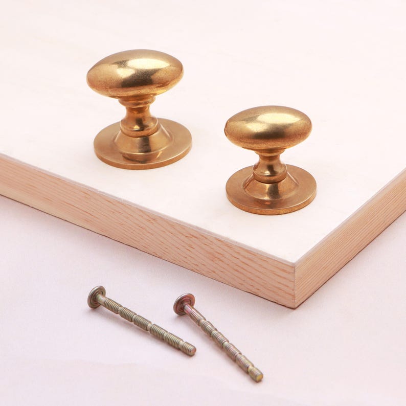 May include: Two polished brass cabinet knobs with oval-shaped heads and circular bases, accompanied by two matching metal mounting screws. The knobs feature a smooth, reflective metallic finish and are designed for attachment to furniture doors.