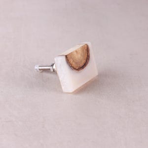 May include: A square cabinet knob featuring a semi-circular slice of natural brown wood embedded within a translucent, off-white resin block. A silver-toned metal bolt with a nut extends from the back of the square-shaped furniture hardware piece.