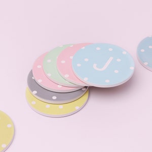 Può includere: Un set di sei sottobicchieri rotondi con un motivo a pois. Il sottobicchiere superiore è blu chiaro con pois bianchi e la lettera "J" in bianco. Gli altri sottobicchieri sono in tonalità di rosa, verde e grigio.