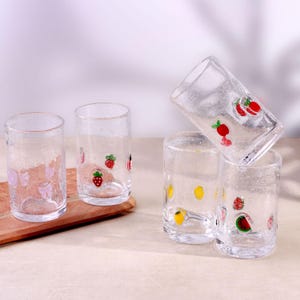 Può includere: Un gruppo di bicchieri trasparenti decorati con motivi di frutta. I bicchieri presentano fragole rosse, limoni gialli e fiocchi rosa. I bicchieri sono disposti su una superficie di legno, con un bicchiere inclinato.
