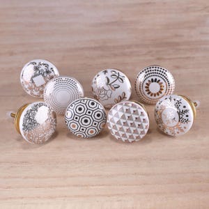 May include: A collection of eight decorative cabinet knobs. Each knob features a white ceramic base with various patterns in gold, black, and gray. Designs include floral, geometric, and dotted patterns. The knobs have gold-colored metal hardware.