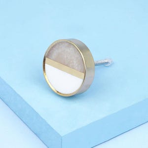 Op de afbeelding: Een ronde kastknop met een goudkleurige metalen rand en een decoratief oppervlak. Het oppervlak is verdeeld in drie secties: een lichtbeige bovenkant, een goudkleurige horizontale streep en een witte onderkant. De knop heeft een zilveren schroef.