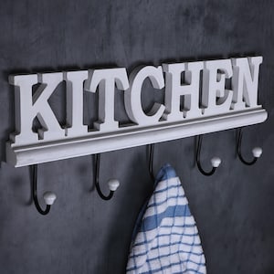 May include: A white wooden sign with the word "KITCHEN" in large letters, above a row of black hooks with white tips. A blue and white checked tea towel hangs from one of the hooks.