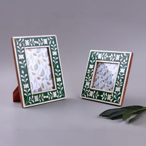 May include: Two decorative photo frames. The frames have a dark green border with white floral accents. The frames are rectangular with a wooden backing and stand. The background of the photo is a leaf pattern.