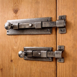 Puede incluir: Dos cerrojos de puerta de metal de estilo antiguo con acabado martillado. Cada cerrojo tiene una base rectangular, una barra deslizante y un pomo redondo. Los cerrojos están montados en una puerta de madera.
