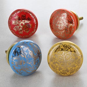 May include: Four decorative cabinet knobs in red, orange, blue, and yellow. Each knob features a gold design. The red knob has an owl pattern, the orange has a floral pattern, the blue has a swirl pattern, and the yellow has a geometric pattern.