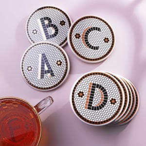 Pode incluir: Um conjunto de bases para copos redondas em cerâmica com um padrão de azulejos hexagonais brancos e as letras A, B, C e D em preto e cobre. Cada base tem um rebordo fino em cobre. Uma caneca de vidro com líquido âmbar está por perto.