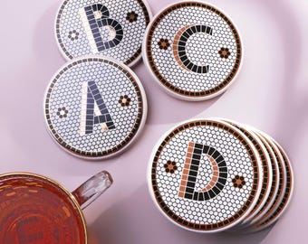 May include: A set of round ceramic coasters featuring a white hexagonal tile pattern with black and copper-colored letters A, B, C, and D. Each coaster is bordered by a thin copper rim. A glass mug filled with amber liquid sits nearby.