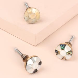 May include: Three decorative cabinet knobs with a mother-of-pearl and abalone shell design. Each knob features a round, segmented face with a gold-coloured base and a silver screw. The knobs are arranged on a light pink surface.