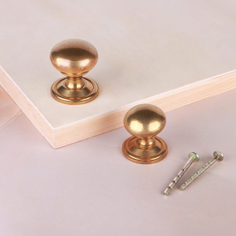 May include: Two round, metallic gold-toned cabinet knobs with a smooth, polished finish and a circular base. Each knob features a central stem connecting the spherical top to the base. Two matching metal mounting screws are included for installation.