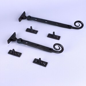 Könnte beinhalten: Zwei schwarze Fensterfeststeller aus Schmiedeeisen mit passender Montagehardware. Jeder Feststeller hat ein dekoratives Scroll-Ende und ein Scharnierdesign. Das Set enthält sechs Montageplatten. Ideal für eine rustikale oder Vintage-Ästhetik.