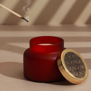 Op de afbeelding: Een rode glazen kaars pot met een gouden deksel met een geometrisch ontwerp. De kaars is gedeeltelijk gesmolten en het deksel is eraf. Een houten lucifer ligt op een oppervlak.