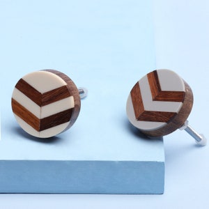 May include: Two round wooden cabinet knobs with a chevron pattern in white and brown. The knobs have a silver metal screw.